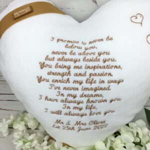 Heart shaped white cushion keepsake with wedding vows embroidered in gold and personalised. To one corner of heart a gold band ribbon and wooden button with just married detail.